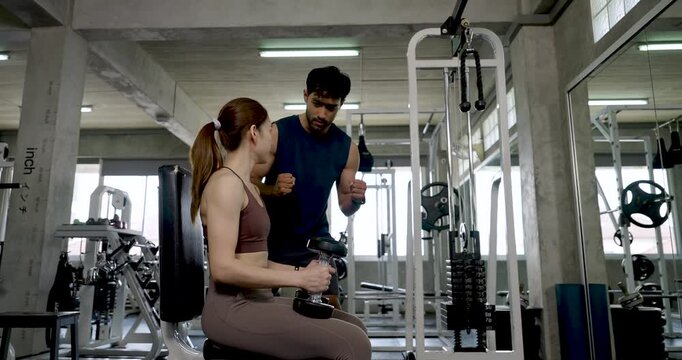 Young Asian couple continuing workout in modern gym, woman bends for dumbbell then sits as man coaches next movement, partner fitness routine with support, arm training, wellness, teamwork goals.
