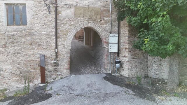 Arco in pietra che introduce un passaggio storico, affiancato da albero e pannello informativo. Atmosfera antica, luminosa e tipica dei borghi europei