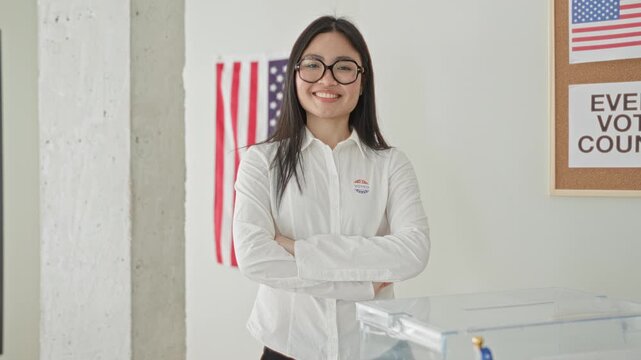 Woman arms crossed by a transparent ballot box with an american flag on the wall in a polling building; civic pride.