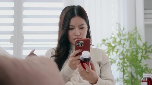 Asian woman holding drug bottle reading information on smartphone at home