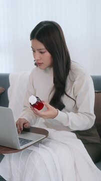 Asian woman holding drug bottle reading information on laptop at home