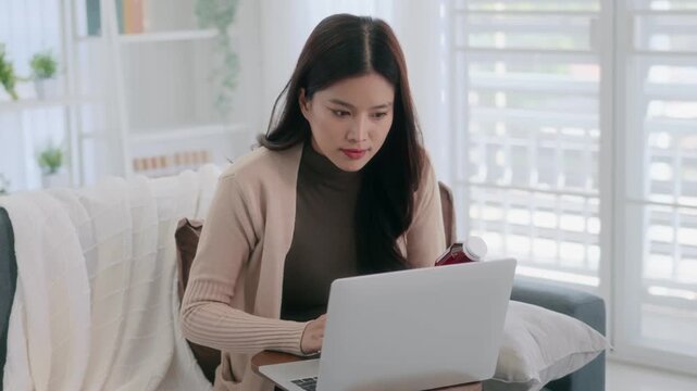 Asian woman holding drug bottle reading information on laptop at home