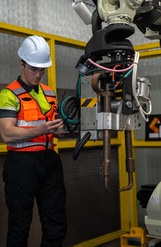 Industrial Robot Technician: A male technician in a hard hat and safety vest supervises an industrial robotic arm in a factory setting, emphasizing precision engineering and automation.