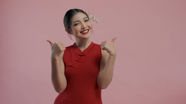 Happy young Asian woman wearing traditional cheongsam qipao dress giving thumb up of approval gesture, isolated by pink background.