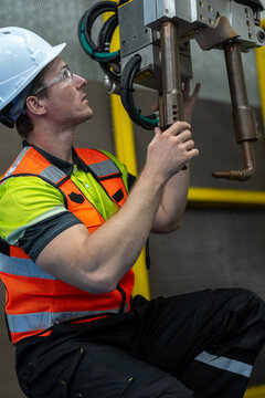Industrial Robot Technician: A male technician in a hard hat and safety vest supervises an industrial robotic arm in a factory setting, emphasizing precision engineering and automation.