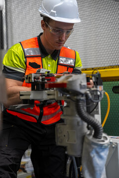 Industrial Robot Technician: A male technician in a hard hat and safety vest supervises an industrial robotic arm in a factory setting, emphasizing precision engineering and automation.