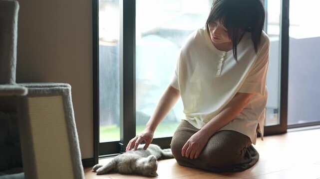 Soft light spills through large windows. Woman kneels, gently touching a resting cat. Her expression shows calm affection. Cat lies peacefully on wooden floor. Quiet moment of domestic tranquility