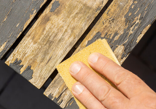 Man sanding loose peeling black paint off a wooden outdoor patio table. Garden DIY chore concept