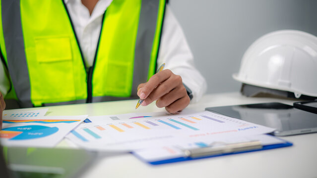 Engineer or project manager holding pen, analyzing yearly summary report chart and financial graphs on office desk with safety hard hat, managing construction project budget.