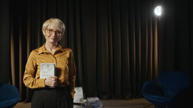 Portrait of middle-aged Caucasian woman holding book and smiling at camera during presentation. Footage is ideal for publishing house promos, author interviews, and educational marketing