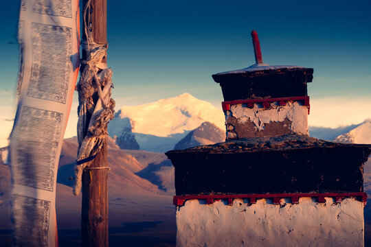 Bathed in the golden glow of sunset, a prayer flag and a shrine stand in quiet reverence at the foot of the Himalayas.