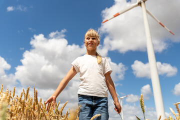 Fototapeta premium Smiling girl walks through golden wheat field under blue sky, with wind turbine and clouds showing clean energy theme. Rural summer scene connects childhood, harvest, nature, sustainability, freedom