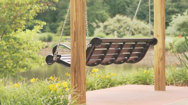 An empty wooden porch swing gently swaying by a peaceful lake in a sunny park