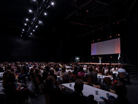 A crowd of attendees fills a big convention hall with a bright stage screen glowing in the dark.