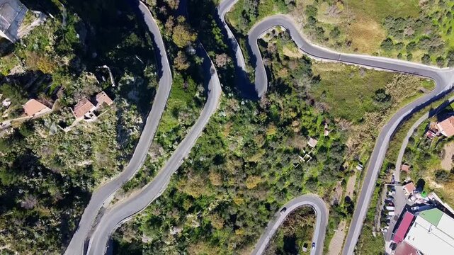 Steep, winding mountain road in eastern Sicily to the medieval hilltop village of Savoca bordered by wild mediterranean vegetation, Drone shot