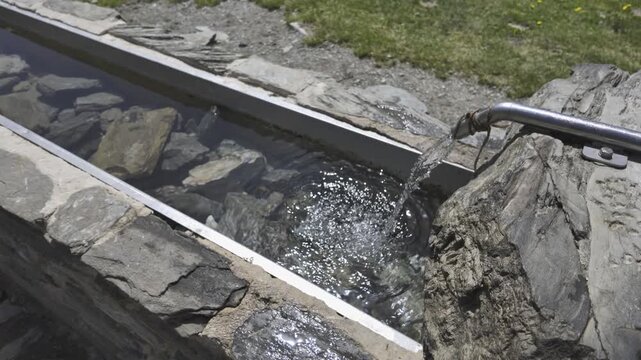 Constant flow of fresh, crystalline drinking water pouring from a metal spout into a long stone waterer for livestock high in the mountains of the Pyrenees, reflecting the bright sunlight