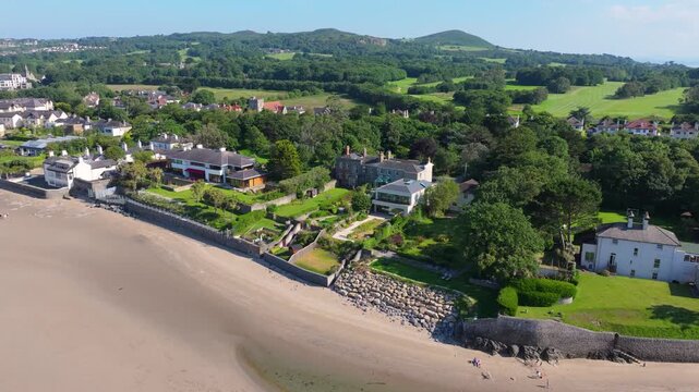 Beachfront villas with landscaped gardens sit above a sandy strand as the drone glides along toward green hills on a sunny day.