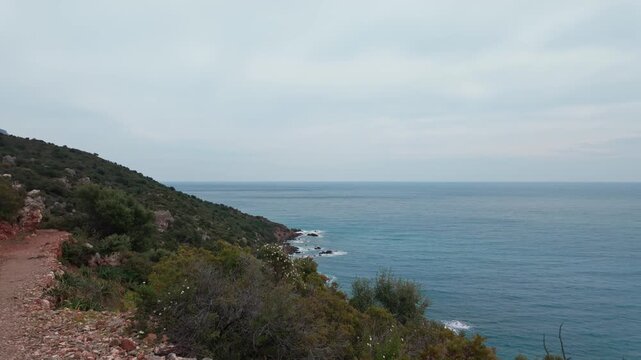 A scenic dirt path winds along a rugged coastline overlooking the turquoise Mediterranean Sea. Cinematic views of the Sardinian coast suitable for travel and wellness content.