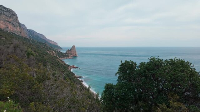 Cinematic aerial sweep of the rugged Pedra Longa coastline in Sardinia. Features limestone cliffs, turquoise Mediterranean waters, and lush green vegetation under a soft sky.