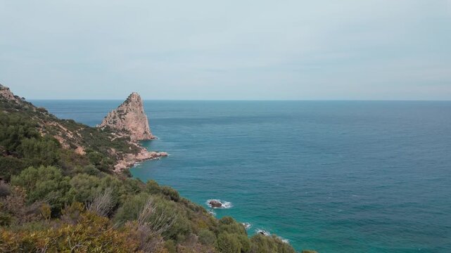 Slow motion aerial reveal of the iconic Pedra Longa limestone needle along the Baunei coast. Cinematic Mediterranean seascape for travel and wellness content.