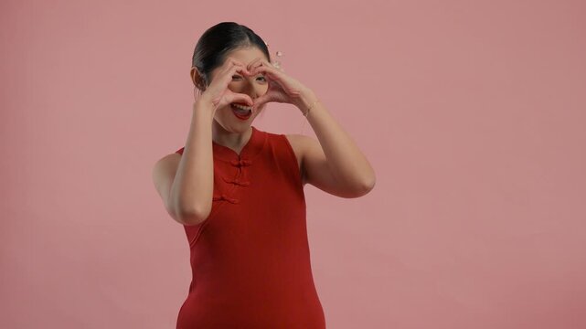 Cute young Asian woman in a traditional cheongsam qipao dress doing love heart finger gesture, isolated by pink background.
