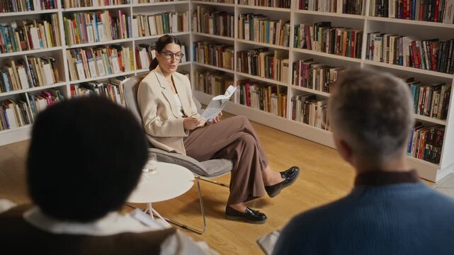 Young adult Caucasian woman wearing glasses reading book to diverse audience in library. Clip is ideal for promoting literary events, educational programs, or community workshops, zoom out