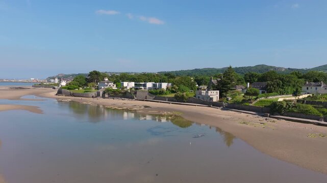 Residential houses overlook a wide sandy inlet at low tide as a drone glides along the quiet shoreline under clear blue skies.