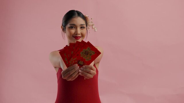 Joyful young Asian woman is holding and giving out angpao or red packet monetary gift, isolated by pink background.