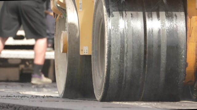 Road roller wheels press hot asphalt. Steam rises from the pavement. Worker's legs stand near the machine. Dust forms around the compactor. Construction work continues efficiently
