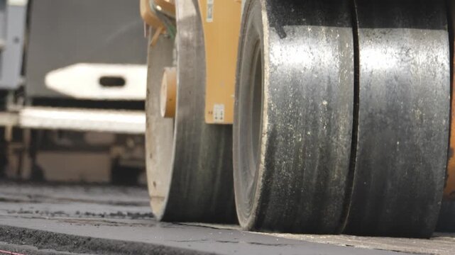 Close-up of the road roller's large tires. The machine compacts fresh asphalt. Dust rises from the surface. No people or animals visible. Ideal for construction projects