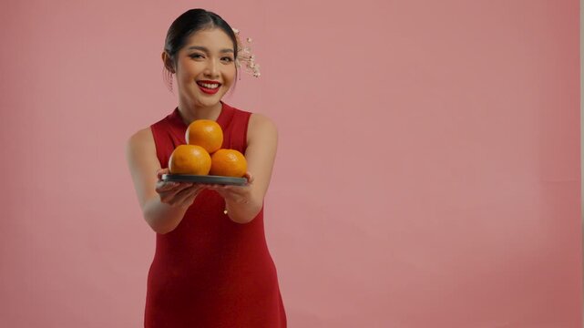 Smiling young Asian woman in a traditional cheongsam dress is giving mandarin oranges, isolated by pink background.