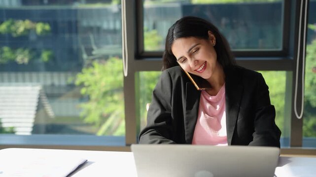 Smiling businesswoman holding smartphone between shoulder and ear while typing on computer, talking with client and managing multiple tasks in modern corporate office workplace.