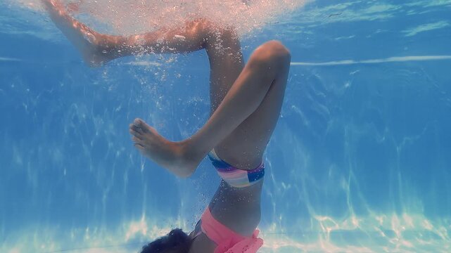 Young girl submerged underwater wearing pink swimwear, swimming in bright blue clear water with sunlight filtering through surface above.