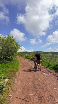 Male cyclist walking bike on sunny rural dirt trail with dramatic clouds and rolling green hills, suitable for outdoor blogs, lifestyle, travel,  Babin Zub Serbia