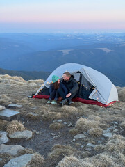 Fototapeta premium Happy couple sitting inside tent at mountain summit during sunset