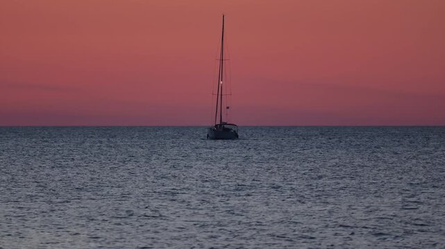Solitary sailboat floating on the calm sea surface under a dramatic red and orange sunset sky with dark storm clouds, Crete