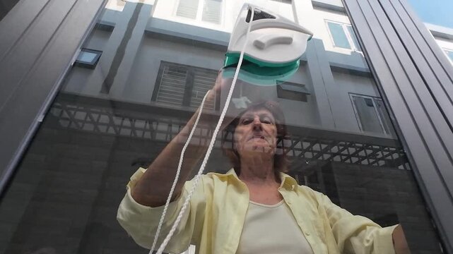 Concentrated woman spends weekend cleaning house with modern magnetic gadget. Female housekeeper works hard, cleaning house windows carefully to achieve spotless glass and perfect domestic order.