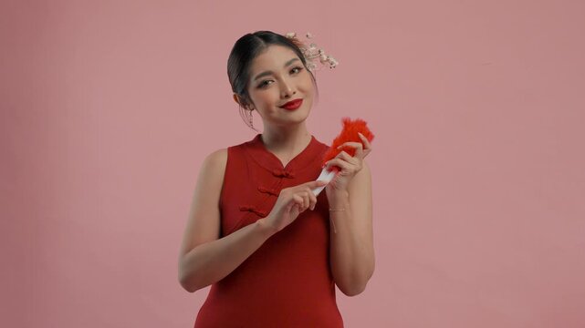 Beautiful young Asian woman in a traditional cheongsam dress posing while holding red feather fan, isolated by pink background.