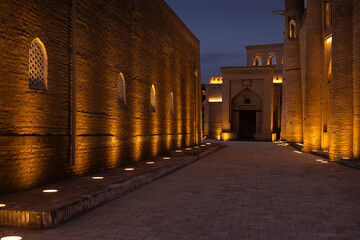 Fototapeta premium Illuminated brick walls and pathway inside Ichan Kala old town in Khiva at twilight. Ancient Islamic architecture with arched windows and warm ground lighting creates a dramatic evening atmosphere.