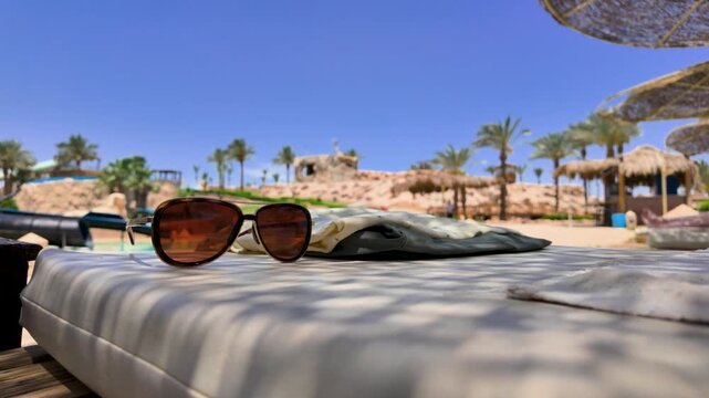 Stylish sunglasses on a cushioned sun lounger at a tropical beach resort with palm trees thatched parasols and blue sky creating a relaxing luxury summer holiday atmosphere for travel themes