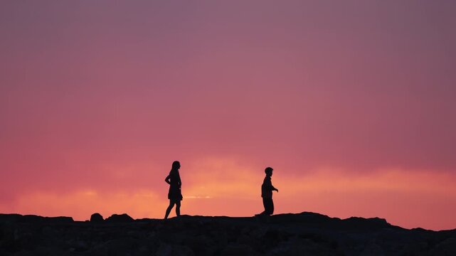 Silhouette of a mother walking with her 11-year-old son wearing an arm brace on a stone pier by the sea against a vibrant pink sunset sky. Inspiring footage of family, recovery, and evening stroll