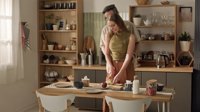 Wide shot of young adult Caucasian woman slicing green apple while middle aged Caucasian man embracing her in kitchen. Footage suitable for healthy lifestyle and relationship marketing campaigns