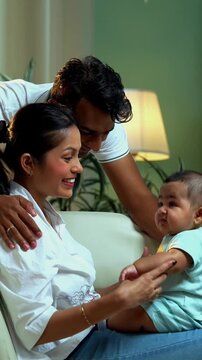 Indian parents catching baby boy while sitting on sofa in modern living room, enjoying fun family bonding, joyful parenthood, love, laughter, care, connection, parenting moments indoors at home