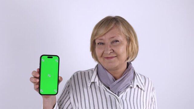 Senior Woman Showing Green Screen Smartphone. Senior woman holding a smartphone with green screen for mobile app, website or online service mockup against a plain light background.