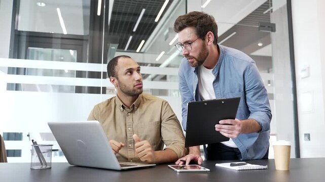Two businessmen discussing startup project details using laptop in business office. Male professional colleagues communicate, exchanging ideas. Manager explain the subordinate. Teamwork, collaboration