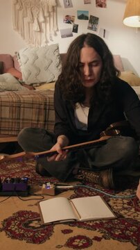 Vertical video of young man playing electric guitar while sitting on floor and writing notes in notebook. Clip for music education or creative hobby marketing campaigns