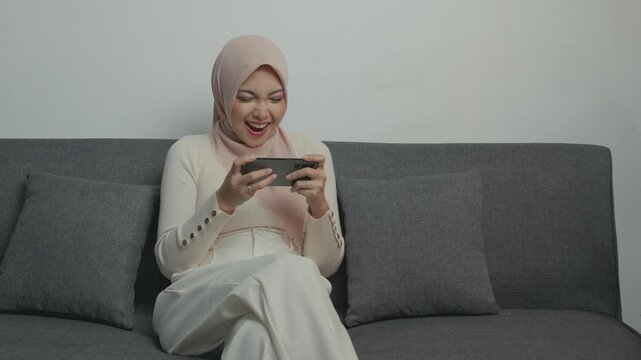 Beautiful Muslim Asian woman in a hijab is using smartphone playing online games while sitting on a sofa.