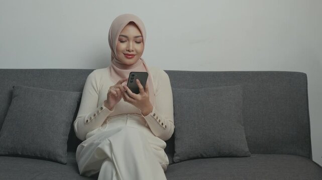 Pretty young Asian Muslim woman in hijab using smartphone while sitting down on a sofa.