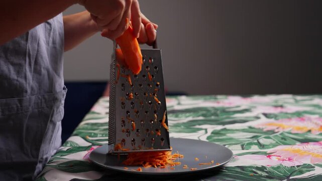 Close up of grating fresh carrot with kitchen grater during homemade cooking using healthy vegetables for preparing delicious meals in home kitchen. High quality 4k footage