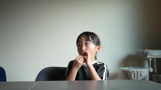 Young child sits quietly at a table. Child holds a small object near their mouth. Child appears thoughtful, eyes gazing forward. Scene suggests a calm, domestic moment